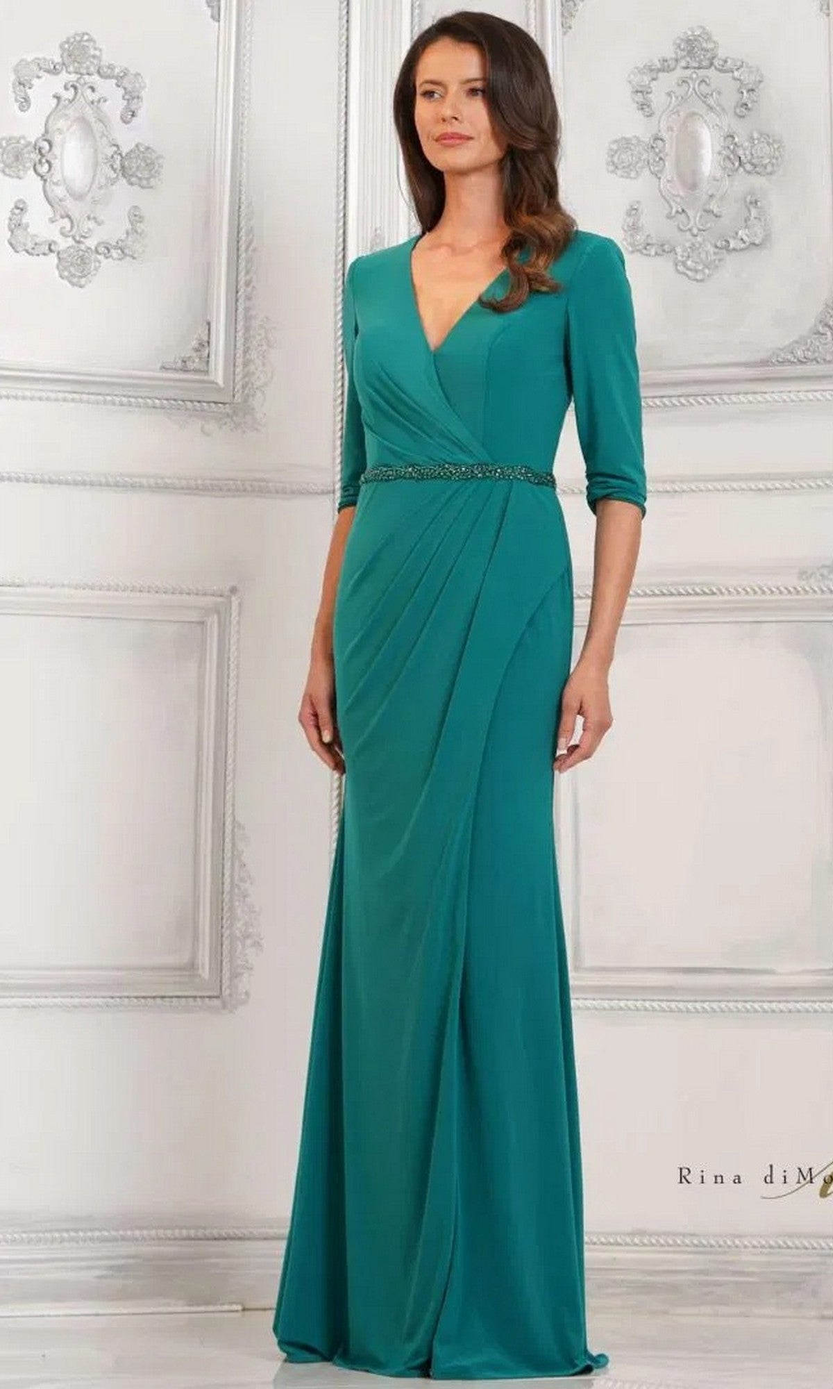 A woman in the Rina Di Montella RD3109 long formal dress, featuring a floor-length emerald matte jersey sheath, three-quarter sleeves, and a beaded waistband, stands against an ornate white background.